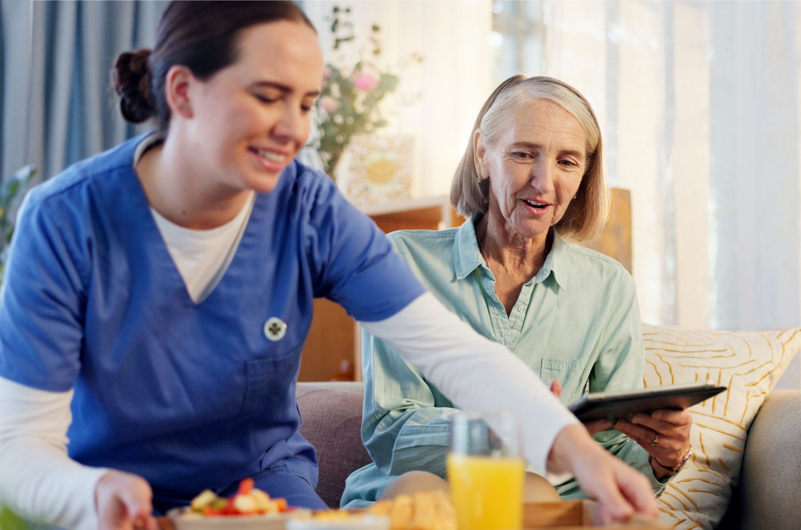 A smiling assisted living caregiver sits beside a resident and places a breakfast tray on the table.