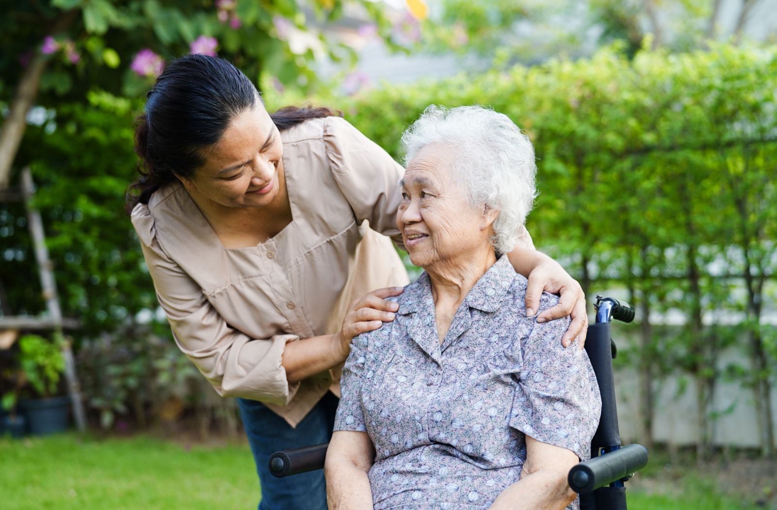 An elderly person sits in a wheelchair and looks up while their younger caregiver holds their shoulders and smiles warmly.