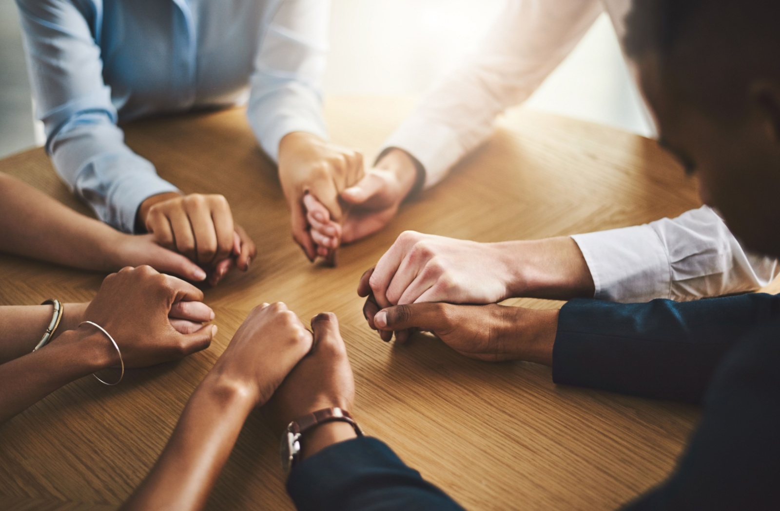 Five sets of hands hold hands tightly on a wooden table in a brightly lit room.