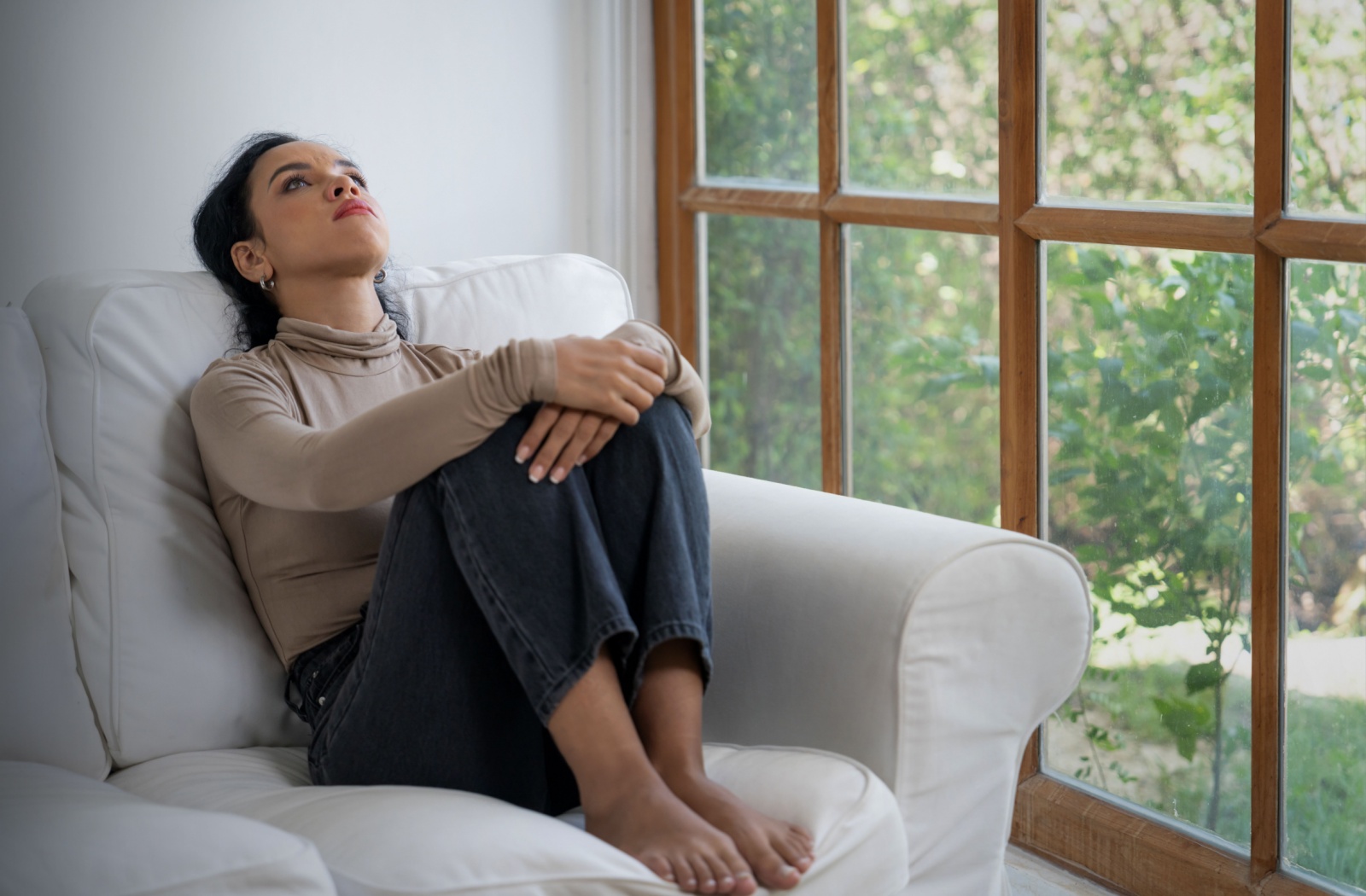 A stressed family caregiver sits alone on their couch next to a window.