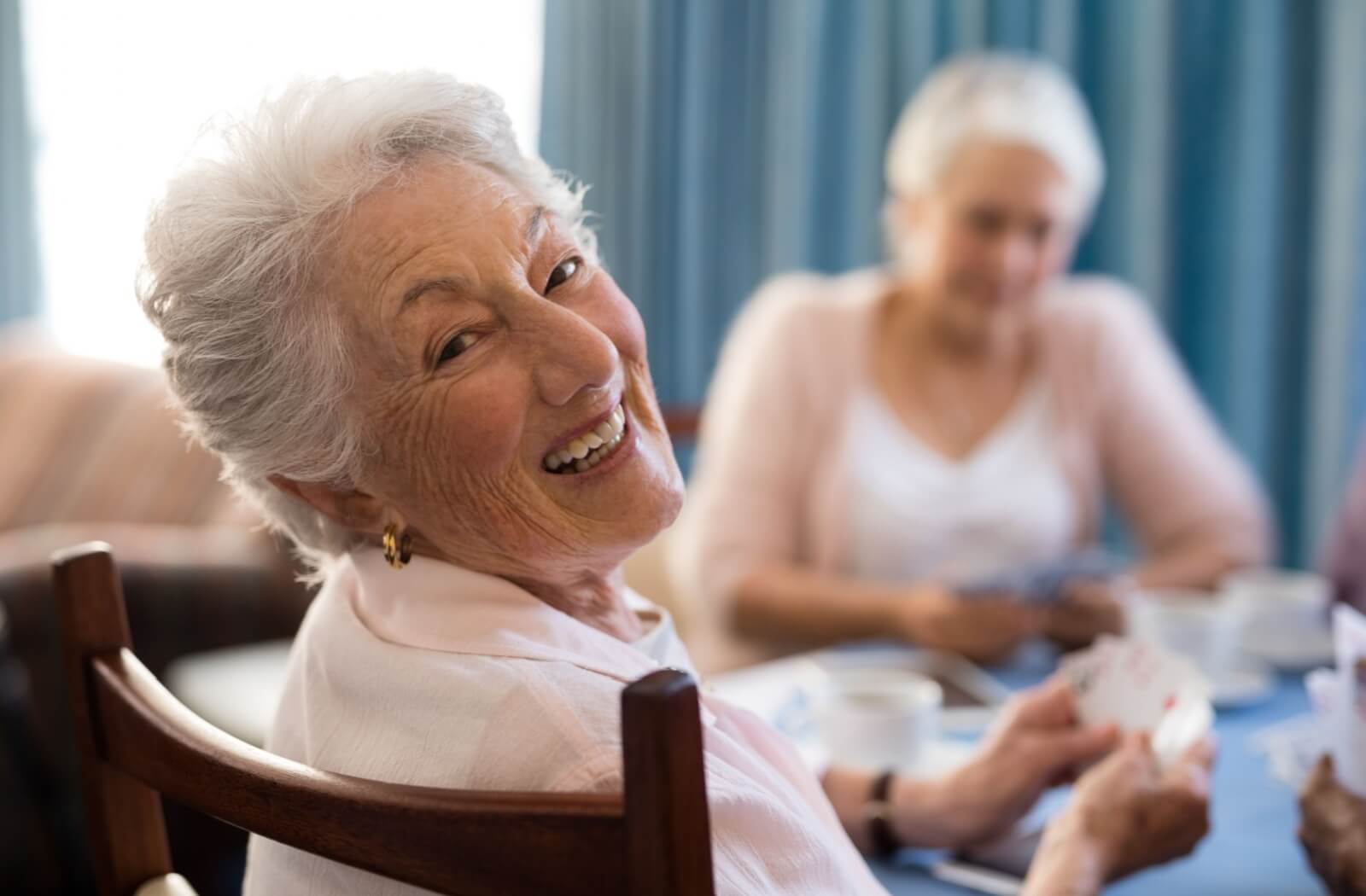 A smiling older adult sitting at a table and enjoying playing cards with other residents in senior living.
