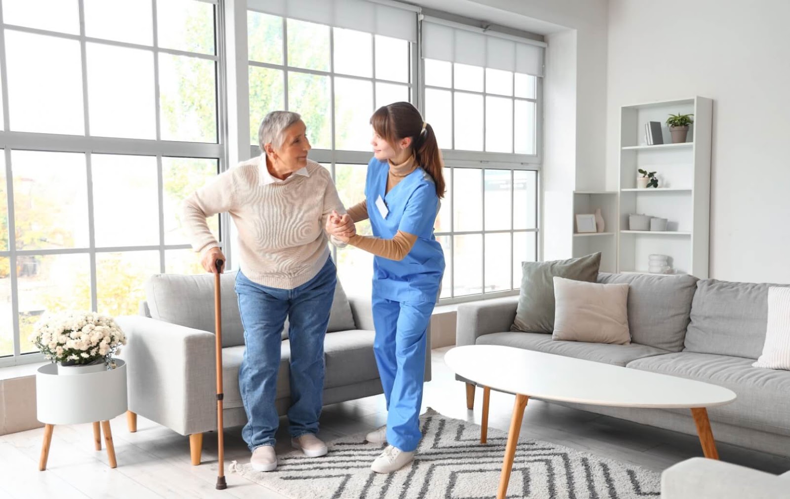 A nurse helps an older adult with a cane stand up from the couch in a cozy, brightly lit living room.
