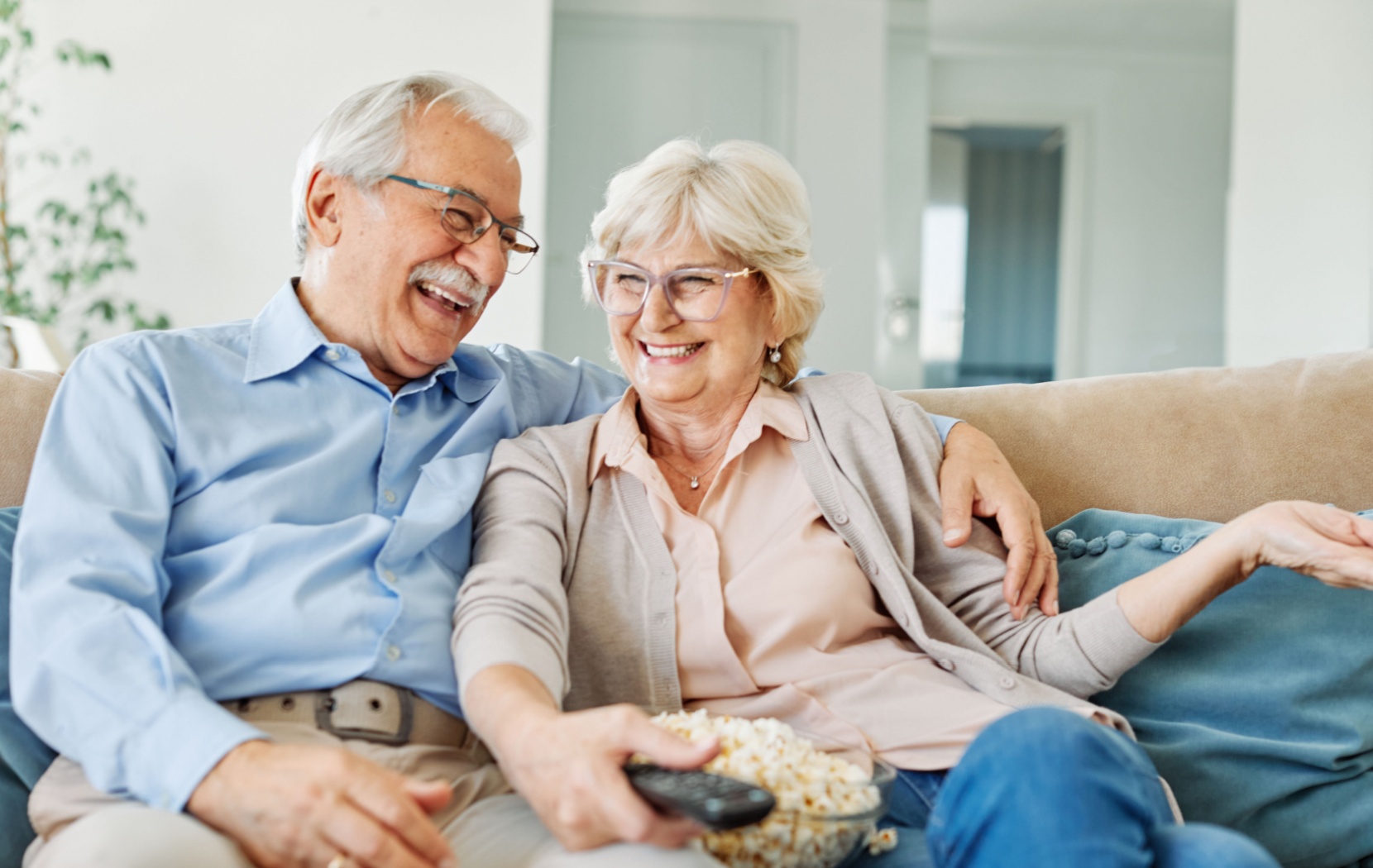 A senior couple sits on their couch in their senior living community. They have a bowl of popcorn and are watching a movie.