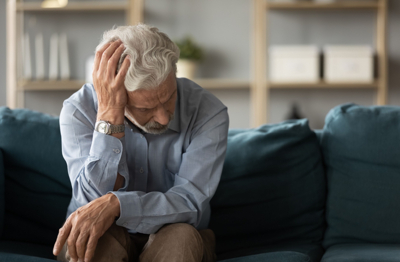 A depressed older adult running a hand through their hair while sitting on their couch in a dark room.