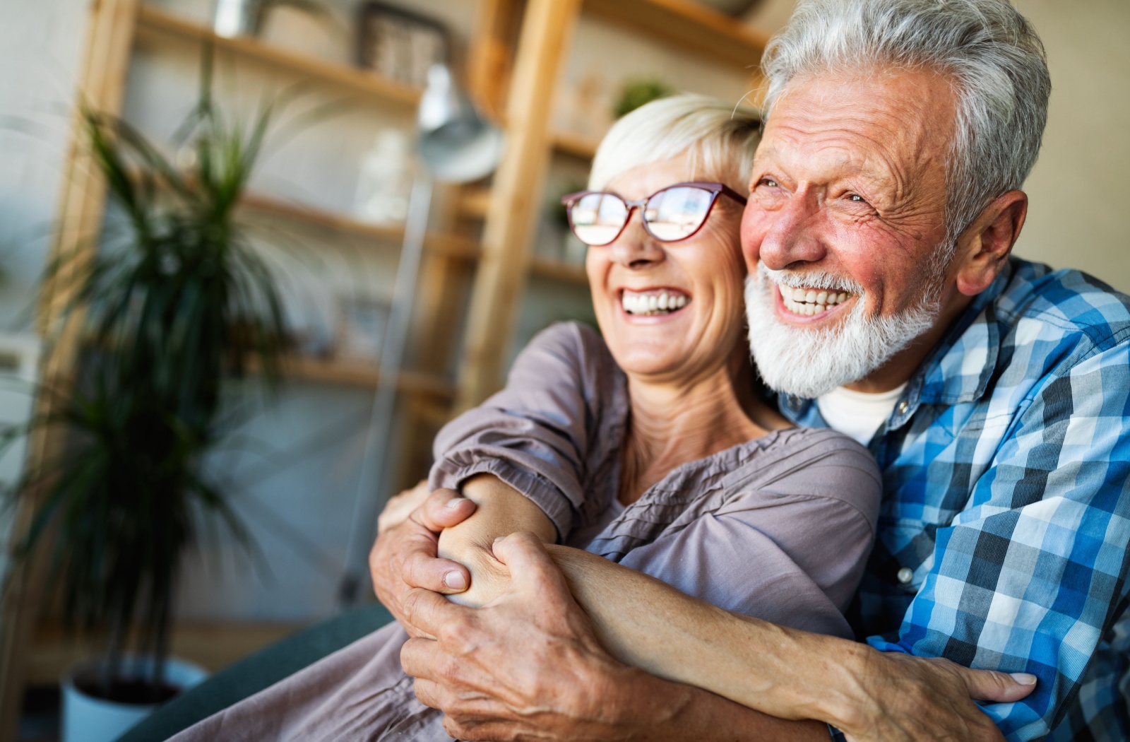 An older adult hugs their smiling spouse from behind while laughing in their new senior living home