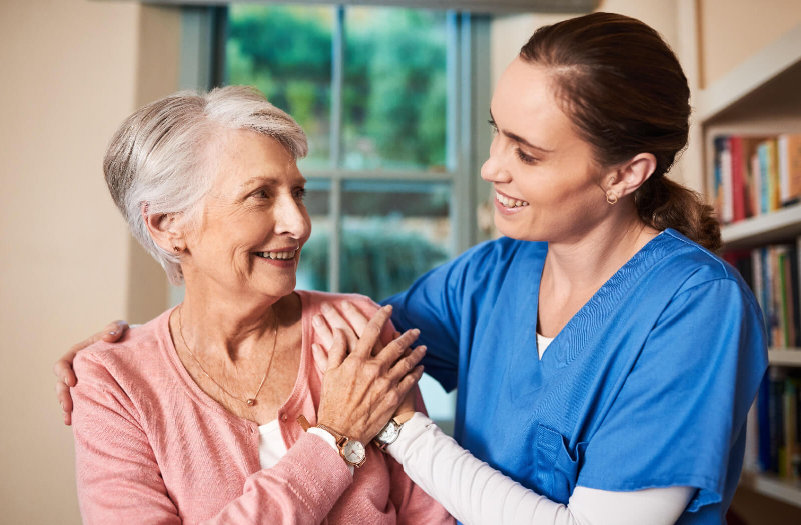 An older adult appreciatively looks at a smiling caregiver with an arm around their shoulder
