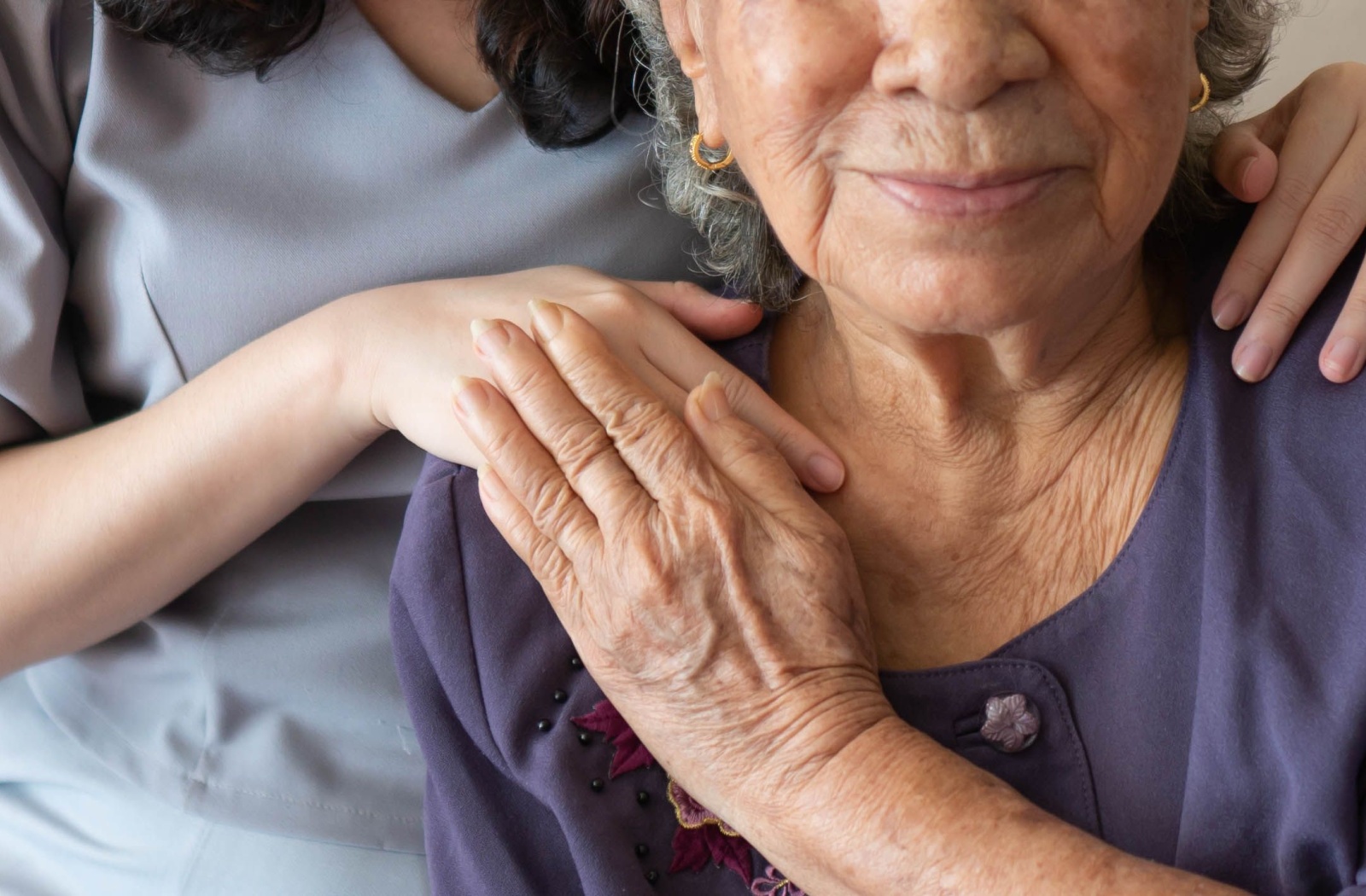 An older adult in assisted living touches the hand of a caregiver behind them while both smile at the camera