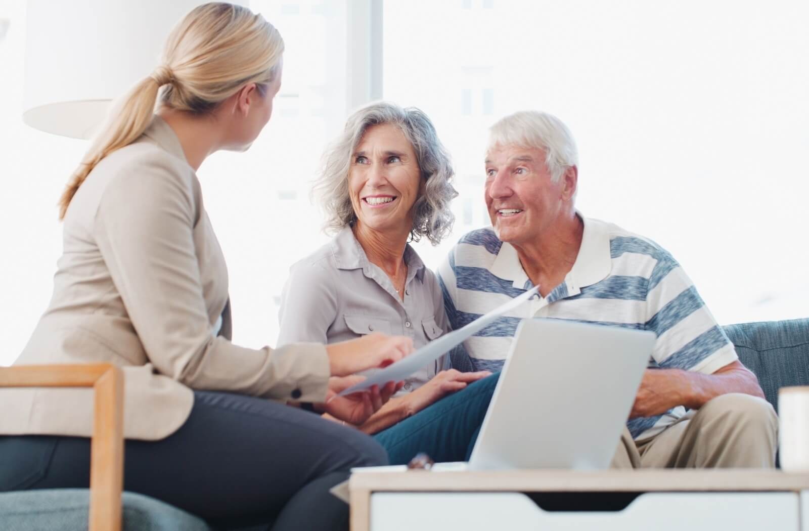 A financial advisor gestures to paperwork in front of an open laptop while helping a smiling senior couple budget for their retirement
