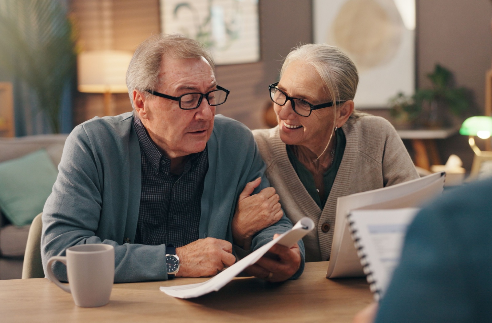 A well-dressed older couple sit arm-in-arm while sipping coffee and reviewing paperwork with an out-of-frame financial advisor for a senior living community