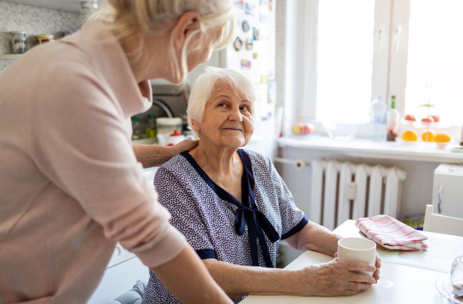 A senior with dementia sitting at a kitchen table looks up at their adult child, who has a hand placed on their shoulder, comforting them.