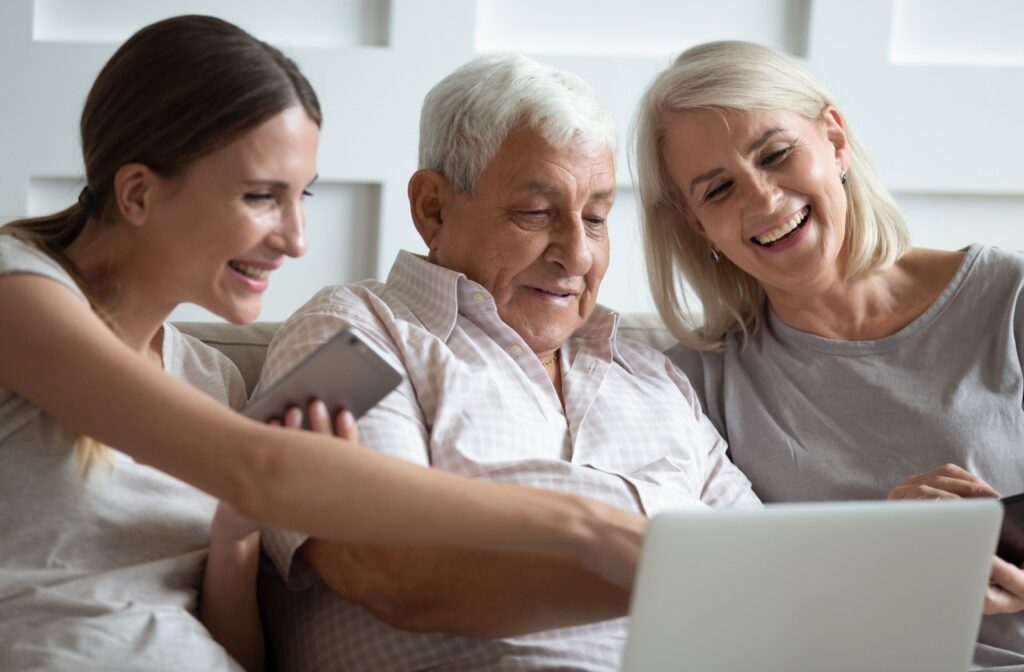 An adult child smiles and points to something on a laptop, while their senior parents watch and laugh